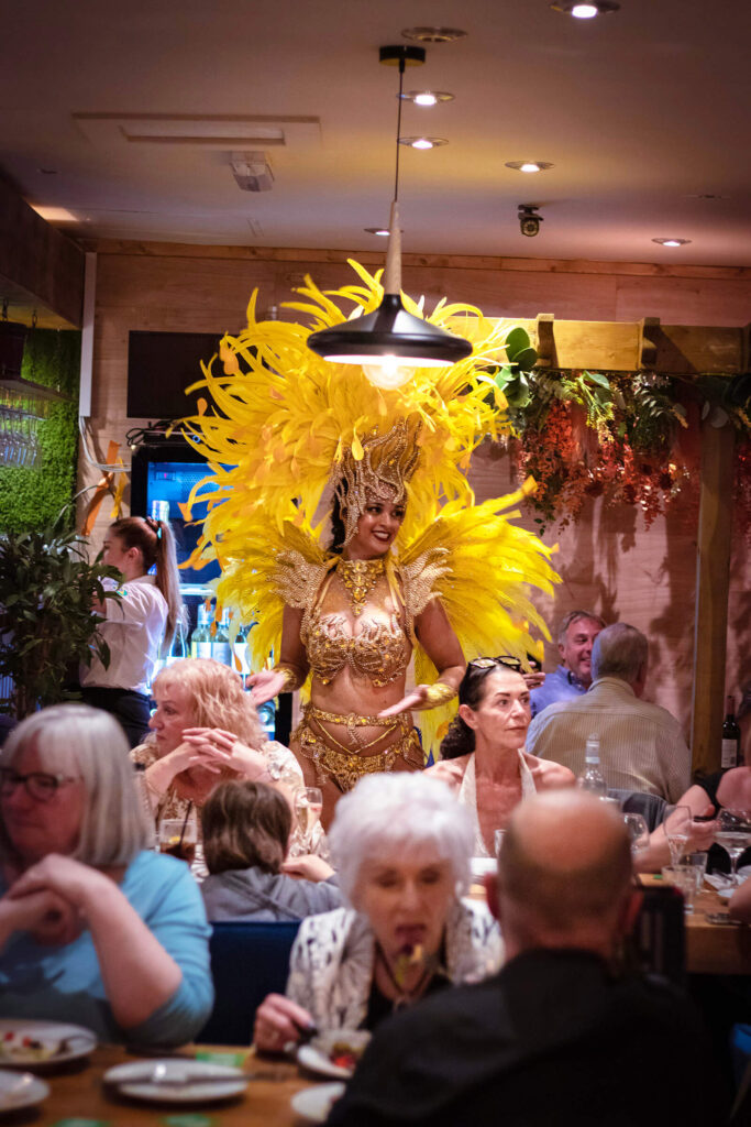 A woman samba dancer in a vibrant yellow costume stands confidently before a gathering of patrons in a bustling restaurant setting.