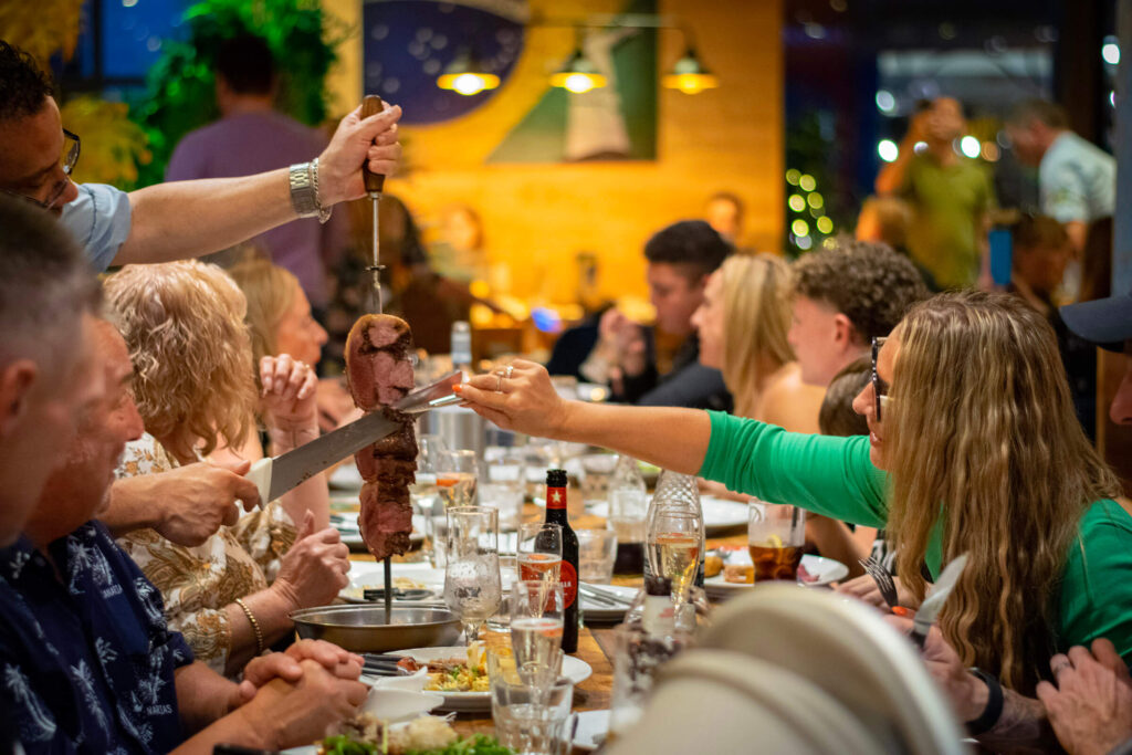 Several people seated at a table, actively discussing, with meat being carved at their table.