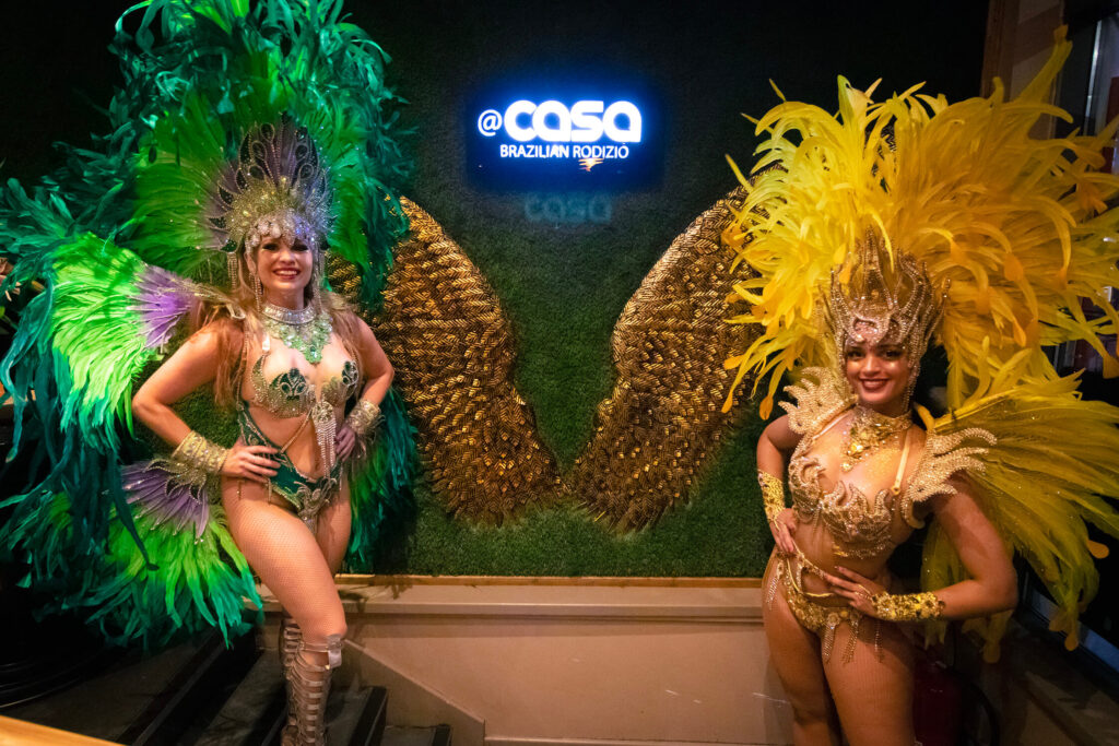 Two women samba dancers in vibrant costumes smile and pose together for a cheerful photo in front of a sign that says Casa Brazilian Rodizio