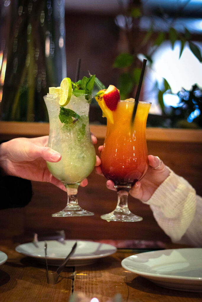 Two people raise their cocktails glasses, a Mojito and tequila sunrise in celebration, toasting over a table filled with beverages.