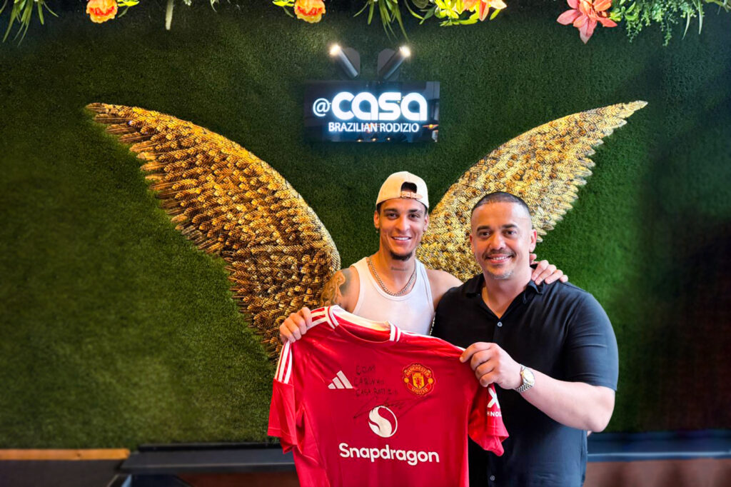 Two men stand in front of a green wall with wings, holding a signed red football jersey. The sign behind them reads Casa Brazilian Rodizio.