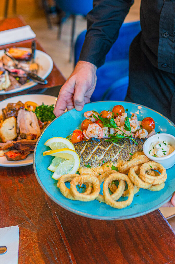 Hands holding a blue plate, ready to serve onto a table. The plate has fish, and seafood on as well as tomatoes and lemon.